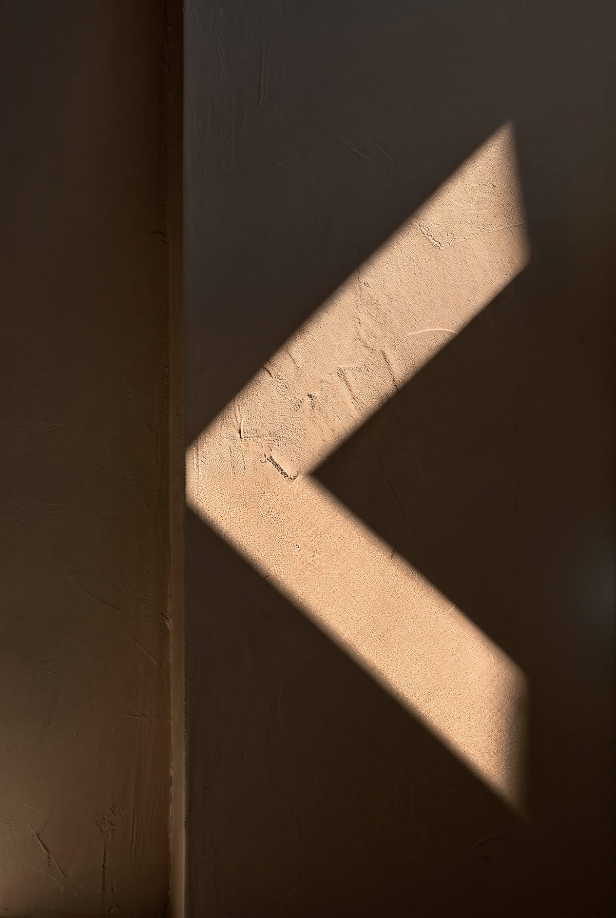 Two bands of late sunlight on a plaster wall meet at the edge of a pillar, forming the letter K.